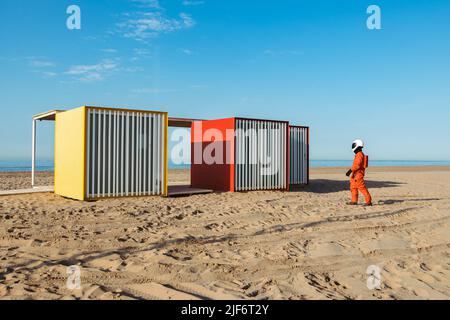 Spaceman in Raumanzug und Helm schlendern in der Nähe kubischer Science-Fiction-Strukturen gegen den blauen Himmel am Sandstrand Stockfoto