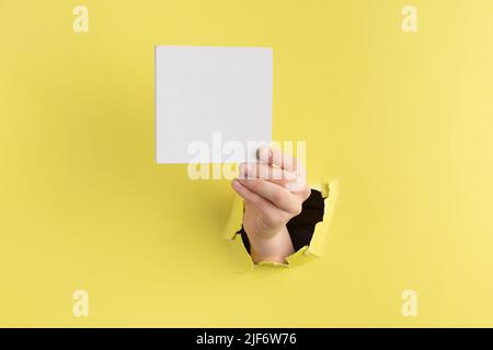 Kind Hand durch Loch in gelbem Papier mit abgerissenen Kanten halten einen weißen quadratischen Holzblock, kopieren Raum Stockfoto