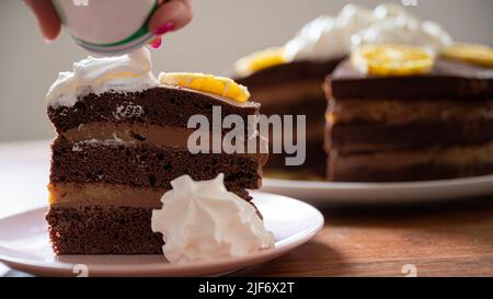 Ein Stück köstlicher veganer Schokoladenorangenkuchen, serviert auf einem Teller mit Schlagsahne und der ganzen Torte im Hintergrund. Stockfoto