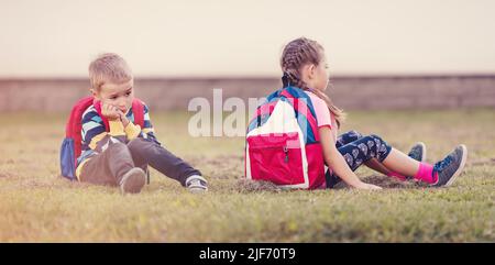 Ein Junge und ein Mädchen streiten sich und sitzen auf dem Gras auf dem Schulhof Rücken an Rücken Stockfoto