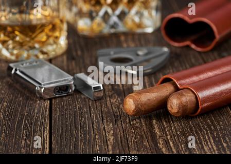 Zwei kubanische Zigarren, ein Cutter und ein Feuerzeug in einem Lederetui auf einem alten braunen Tisch. Glas Whiskey auf unscharfem Hintergrund. Stockfoto