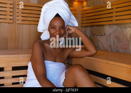 Porträt einer selbstbewussten jungen afroamerikanischen Frau, eingewickelt in Handtücher, die in der Sauna sitzen Stockfoto