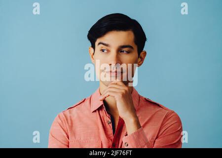 Kopfschuss nachdenklich schöner junger Erwachsener Latino Mann mit Hand auf dem Kinn schwarzen Haaren und rosa Hemd über blauem Hintergrund Blick weg auf Copy Space Studio aufgenommen Stockfoto