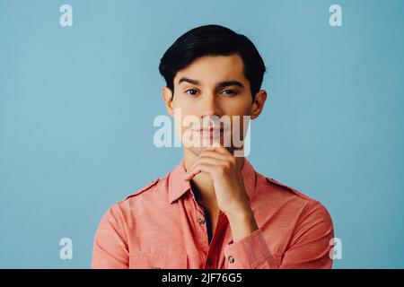 Headshot nachdenklicher, hübscher, junger, erwachsener Latino-Mann mit Hand am Kinn, schwarzen Haaren und pinkem Hemd auf blauem Hintergrund, der auf das Kamerastudio blickt Stockfoto