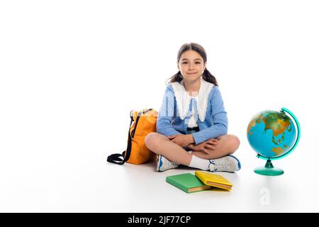 Lächelnder Schüler, der neben Büchern und Globus auf weißem Hintergrund sitzt Stockfoto