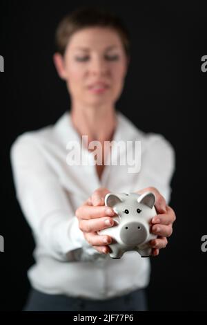 Weißes Schweinebank aus Porzellan, das von einer Frau in Geschäftskleidung, weißem Hemd und grauem Rock auf schwarzem Hintergrund gehalten wird. Konzept für Geld, Finanzen, Einsparungen Stockfoto