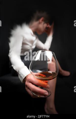 Trinkglas gefüllt mit Alkohol, das von einer Frau in Geschäftskleidung gehalten wird, die mit gekreuzten Beinen und dem Kopf in der Hand sitzt. Das Bild der Frau verschwimmt, als ob sie es wäre Stockfoto