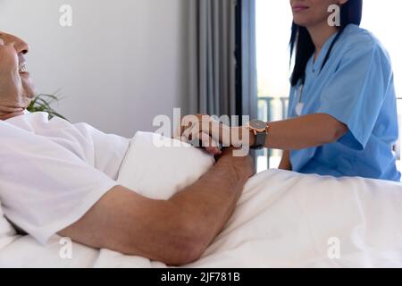 Mittlerer Abschnitt einer birazialen Gesundheitshelferin, die einen älteren kaukasischen Mann auf dem Bett unterstützt Stockfoto