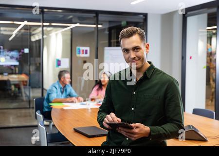 Porträt eines selbstbewussten jungen kaukasischen Geschäftsmannes mit Tablet-pc, während Kollegen im Hintergrund Stockfoto