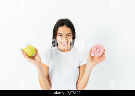 Konzept der gesunden und ungesunden Ernährung. Glückliche junge Frau mit grünem Apfel und Donut, die vor dem weißen Studiohintergrund steht Stockfoto