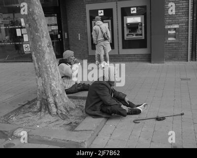 Zwei ältere Obdachlose auf einer Straße, die auf dem Bürgersteig sitzen Stockfoto