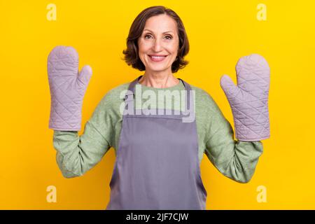 Porträt von attraktiven fröhlich grauhaarige Frau trägt Handschuhe Kochkunst Küche isoliert über hellen gelben Hintergrund Stockfoto