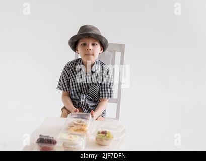 Ein kleiner Junge in einem Hemd und Hut sitzt vor Kuchen mit einem Telefon in den Händen. Stockfoto