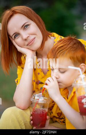 Der rothaarige junge Junge und die Mutter trinken rote Limonade. Sommer Erholung im Freien. Stockfoto