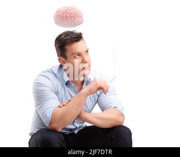 Nervöser junger Mann, der eine Zigarette raucht und ein Gehirn über seinem Kopf schwebt, isoliert auf weißem Hintergrund Stockfoto