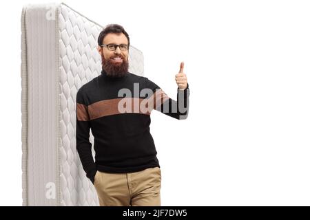 Lächelnder bärtiger Kerl, der Daumen hoch zeigt und sich auf eine isolierte Matratze auf weißem Hintergrund lehnt Stockfoto