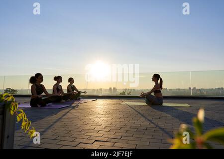 Multiethnische junge Frauen meditieren mit Yoga-Lehrerin während der Gruppenklasse im Freien. Stockfoto