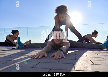 Afrikanischer Ausbilder hilft flexiblen kaukasischen Frau bei Gruppen-Yoga-Kurs im Freien. Stockfoto