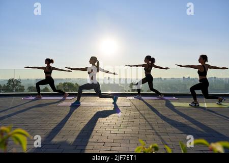 Weibliche Silhouetten stehen in Yoga-Kriegerpose bei der Gruppenklasse im Freien bei Sonnenaufgang. Stockfoto