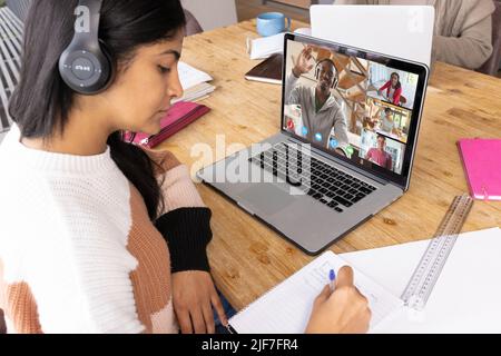 Biracial fokussiertes Teenager-Mädchen, das Kopfhörer trägt und Notizen im Buch schreibt, während es an einem Online-Kurs teilnimmt Stockfoto