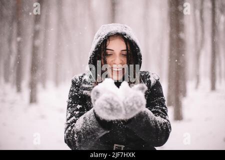 Glückliche lächelnde junge Frau, die während des Schneefalls im Winter Schnee hält Stockfoto
