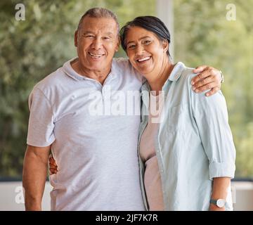 Porträt eines glücklichen älteren Paares, das lächelt und zu Hause vor einem Fenster steht und die Kamera anschaut. Reife hispanische Mann und Frau teilen romantisch Stockfoto