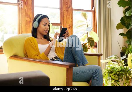 Frau, die ihr Smartphone benutzt und Musik über Kopfhörer hört, während sie sich in einem hellen Wohnzimmer wohlfühlt und entspannt. Eine junge Frau, die auf einem sitzt Stockfoto