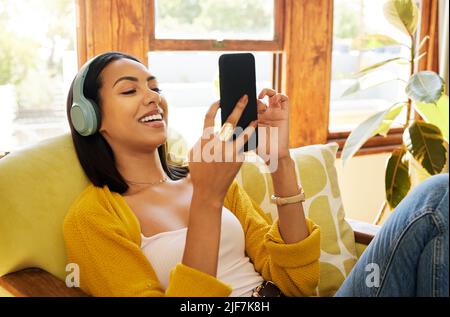 Hispanische Frau, die ihr Smartphone benutzt und Musik über Kopfhörer hört, während sie sich in einem hellen Wohnzimmer wohlfühlt und entspannt. Eine junge Frau Stockfoto