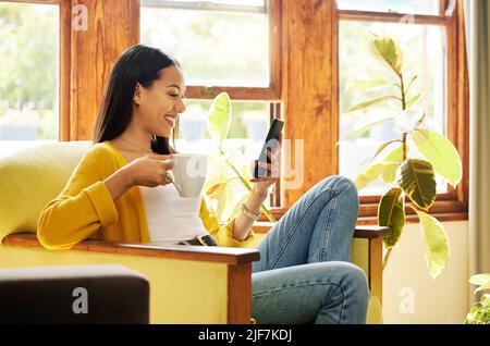 Hispanische Frau, die ihr Smartphone benutzt und Kaffee trinkt, sitzt vor einem Fenster in einem hellen Wohnzimmer. Seitenprofil einer jungen Frau, die aussieht Stockfoto