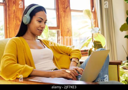 Hispanische Frau tippt auf einem Laptop und hört Musik über Kopfhörer, lächelt und arbeitet in einem hellen Wohnzimmer. Eine junge Frau, die auf einem Stuhl sitzt Stockfoto