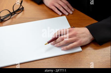 Frau, die einen Stift auf einem leeren weißen Papier hält, oben aus der Nähe, Hintergrund des Bürotisches aus Holz. Weibliche linke Hand unterzeichnet auf leerem Dokument, kopieren Sie den Freitext Stockfoto