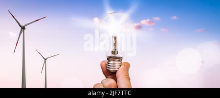 Hand hält eine Glühbirne neben zwei Windturbinen vor einem sonnigen blauen Himmel Stockfoto