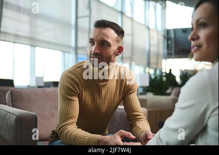 Konzentrieren Sie sich auf einen gutaussehenden arabischen Mann im Pullover, der wegschaut, die Hände seiner Freundin hält und in der VIP-Lounge des International airpo auf einem Stuhl sitzt Stockfoto