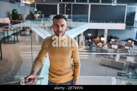 Ein gutaussehender, selbstbewusster junger Mann aus dem Nahen Osten blickt selbstbewusst in die Kamera, während er in der VIP-Lounge des internationalen Flughafens die Treppe hinaufgeht Stockfoto