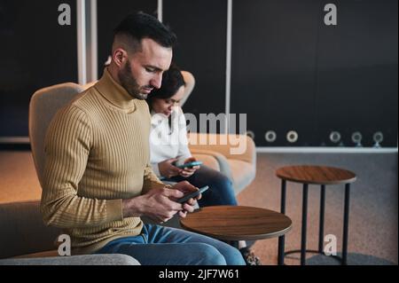 Konzentriertes Multitasking des jungen kaukasischen Mannes mit dem Mobiltelefon, Hotelbuchung, Sitzgelegenheit neben seiner Partnerin, Freundin, Frau in der VIP-Loun Stockfoto