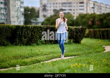 Morgenlauf im öffentlichen Park in der Nähe von Wohngebieten der Stadt im Frühling durch schlanke Frau in Sportkleidung. Stockfoto