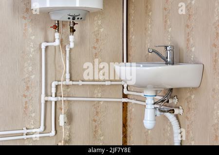 Sanitär-Pipeline der Hauswasserversorgung mit elektrischem Warmwasserspeicher und Waschbecken im Landhaus. Stockfoto