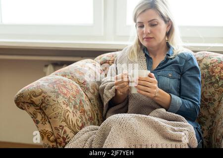 Nahaufnahme der Hände mit einer blauen Tasse Tee oder Kaffee. Modell und kuschelig kariert sitzt, entspannen Sie sich zu Hause auf dem Sofa Stockfoto