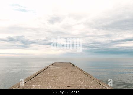 Leerer Betonsteg zum Meer mit dramatischem Himmel und ruhigem Wasser, verlassene industrielle Anlegestelle Stockfoto
