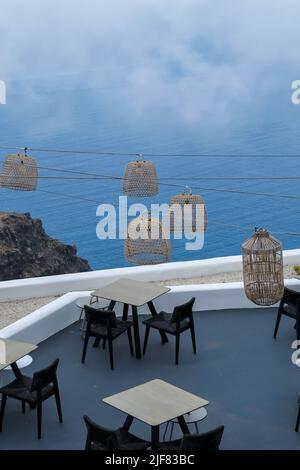 Santorini, Griechenland - 13. Mai 2021 : Blick auf eine malerische Terrasse mit Tischen, Stühlen und Lichtern mit Blick auf einen riesigen Felsen und die Ägäis Stockfoto