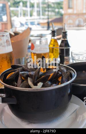 Gekochte Muscheln in einer schwarzen Schüssel und Bier in einem Restaurant. Meeresfrüchte. Klassische traditionelle französische Küche. Leckeres Abendessen. Stockfoto