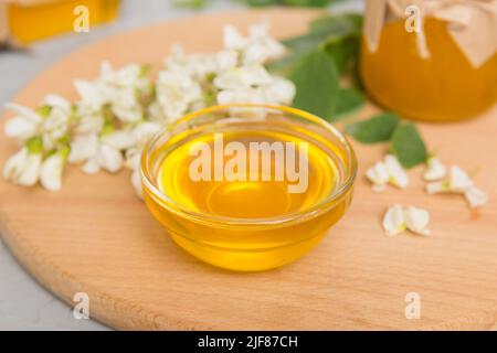 Süßes Honigglas umgab die Akazienblüten des Frühlings. Honig fließt aus einem Löffel in ein Glas. Gläser von klarem frischen Akazienhonig auf Holzgrund. Stockfoto