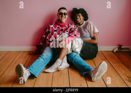 Fröhliche Hipster-Frau und schwuler Mann, die zu Hause auf dem Hartholzboden gegen die rosafarbene Wand sitzen Stockfoto