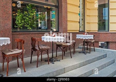 Holzstühle und runde Tische mit weißen Tischdecken auf der Sommerterrasse aus Granit in der Nähe des Cafés oder Restaurants Stockfoto