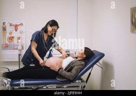 Lächelnde Ärztin, die den Bauch einer Schwangeren während der Untersuchung in einer medizinischen Klinik misst Stockfoto