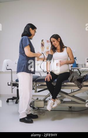 Die ganze Länge der Ärztin überprüft den Blutdruck einer Schwangeren, die auf einer Liege in einer medizinischen Klinik sitzt Stockfoto