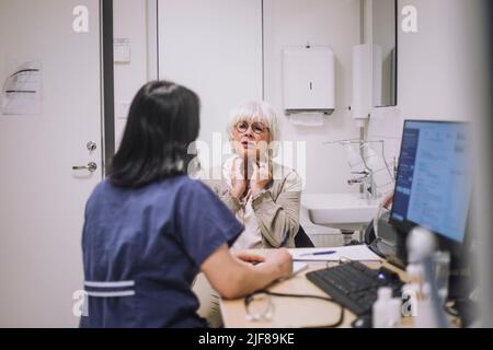Ältere Frau erklärt dem Otolaryngologen, der in der medizinischen Klinik am Schreibtisch sitzt, das Rachenproblem Stockfoto