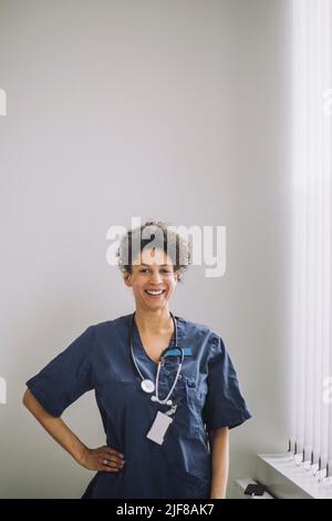 Porträt einer glücklichen Ärztin, die im Krankenhaus mit der Hand auf der Hüfte an der weißen Wand steht Stockfoto