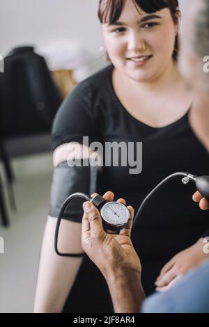 Lächelnde Patientin, die einen Arzt ansieht, der den Blutdruck in einer medizinischen Klinik untersucht Stockfoto