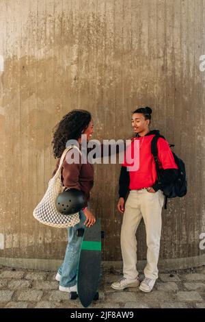 Mann mit der Hand in der Tasche, der mit einer Freundin spricht, die Skateboard hält, während er gegen die Wand steht Stockfoto